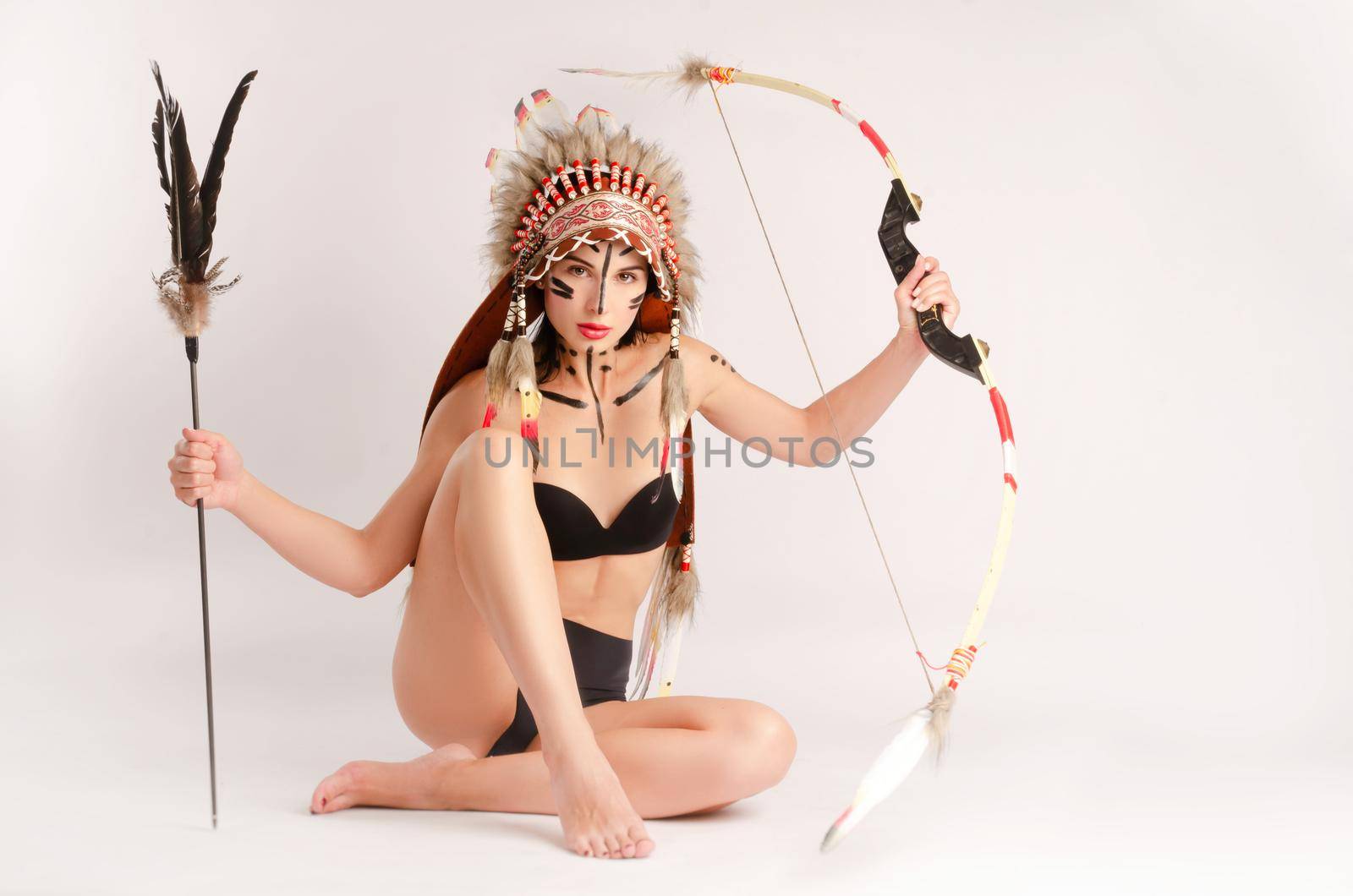 a woman in the image of indigenous peoples of America with a bow and arrow poses sitting on a light background by Rotozey