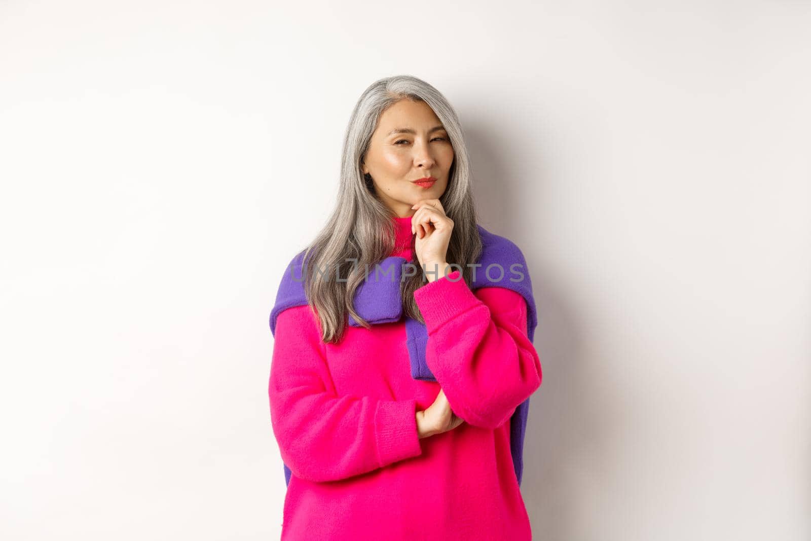 Image of thoughtful asian senior woman squinting suspicious at camera, having assumption, standing over white background and thinking by Benzoix