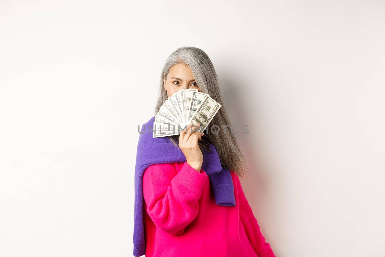 Shopping concept. Rich and fashionable korean senior woman hiding face behind money, looking at camera, standing over white background.