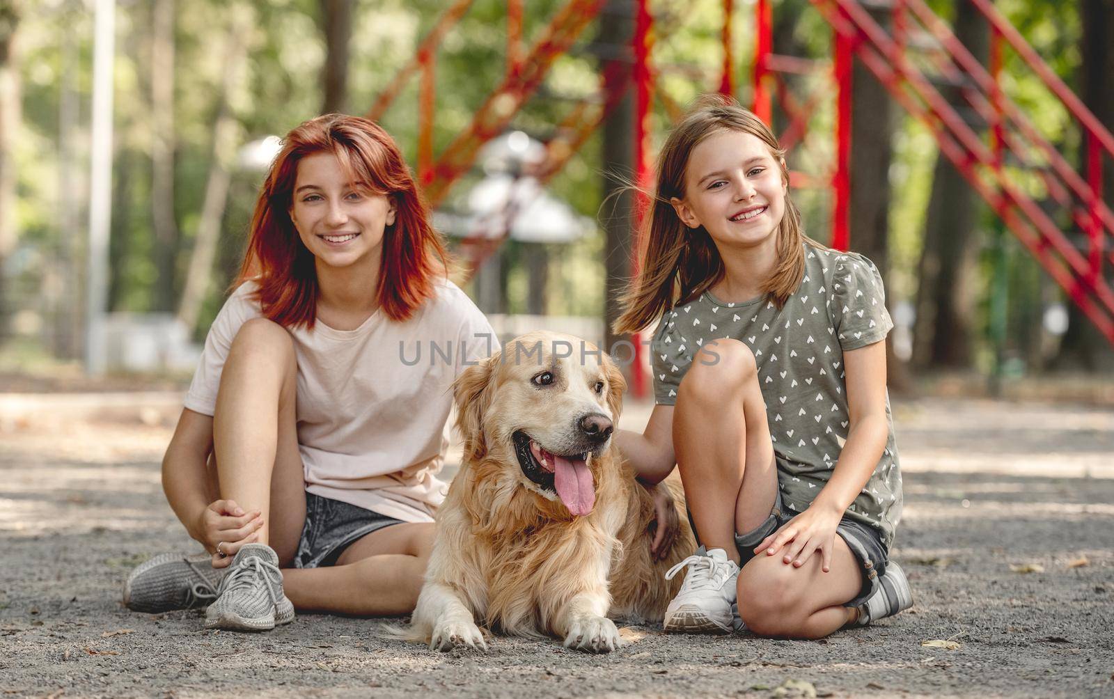 Beautiful girls with golden retriever dog sitting in the park and smiling. Sisters with doggy pet outdoors