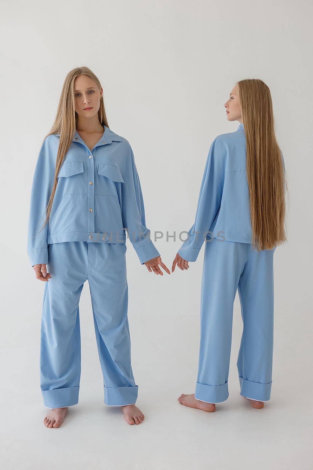 two young twin sisters with long blond hair posing on white background in oversize clothes with bare feet. They wear blue suits that look like pajamas and pull hands to each other but do not touch