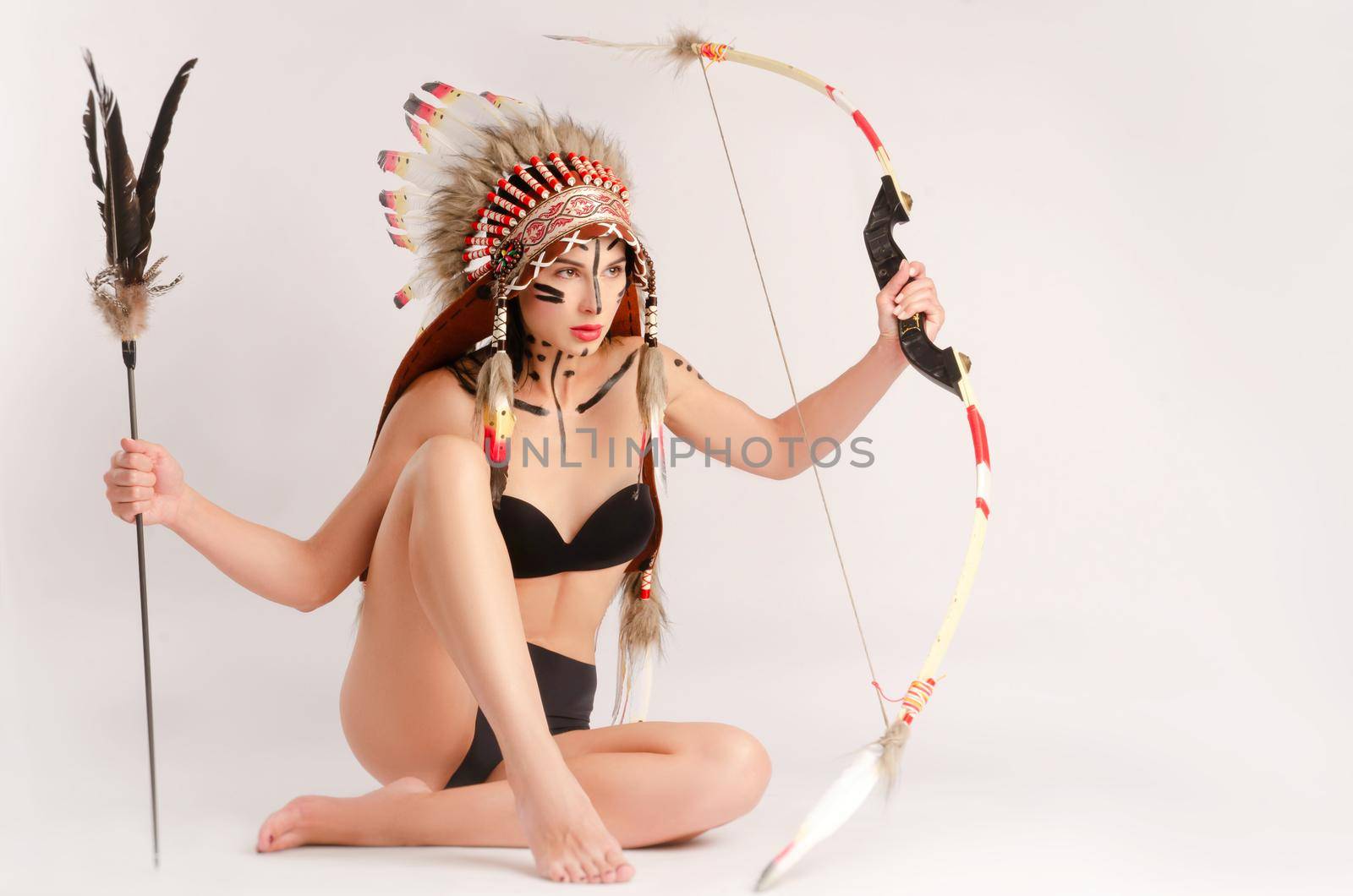 the woman in the image of indigenous peoples of America with a bow and arrow poses sitting on a light background