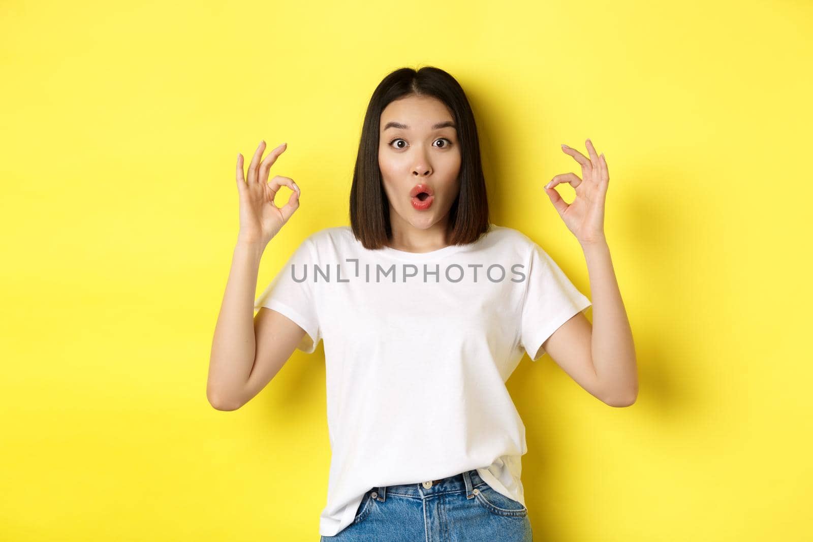 Impressed korean girl saying WOW, showing okay signs and looking amazed, standing against yellow background.