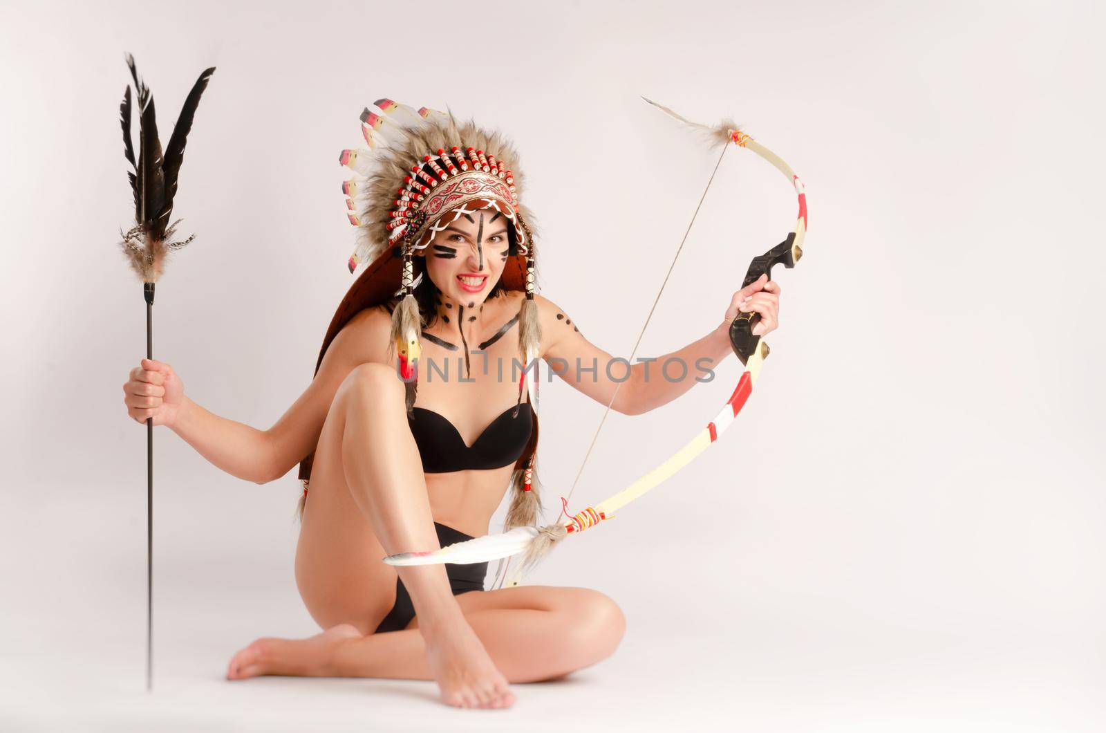 the woman in the image of indigenous peoples of America with a bow and arrow poses sitting on a light background