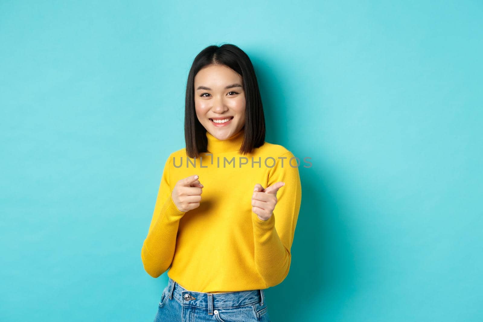 Smiling young asian woman pointing fingers at camera, choosing you, inviting to event, standing happy against blue background.