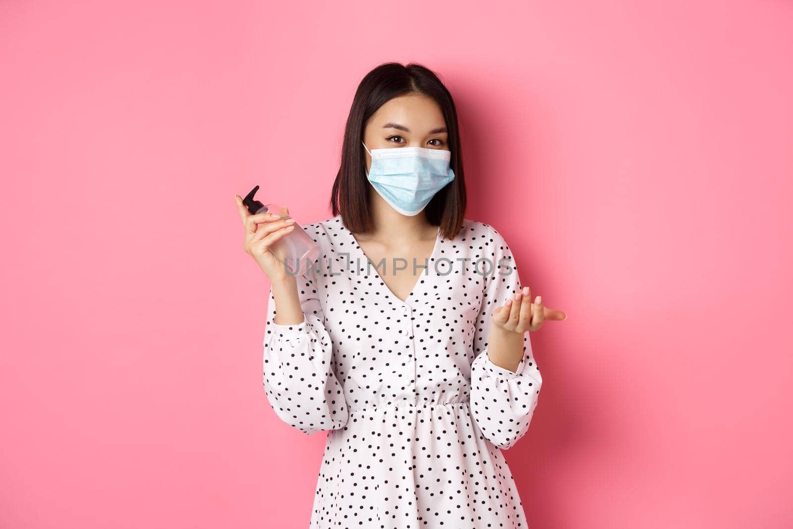 Covid-19, pandemic and lifestyle concept. Cute asian woman clean hands with sanitizer, using antiseptic and wearing face mask, standing over pink background.