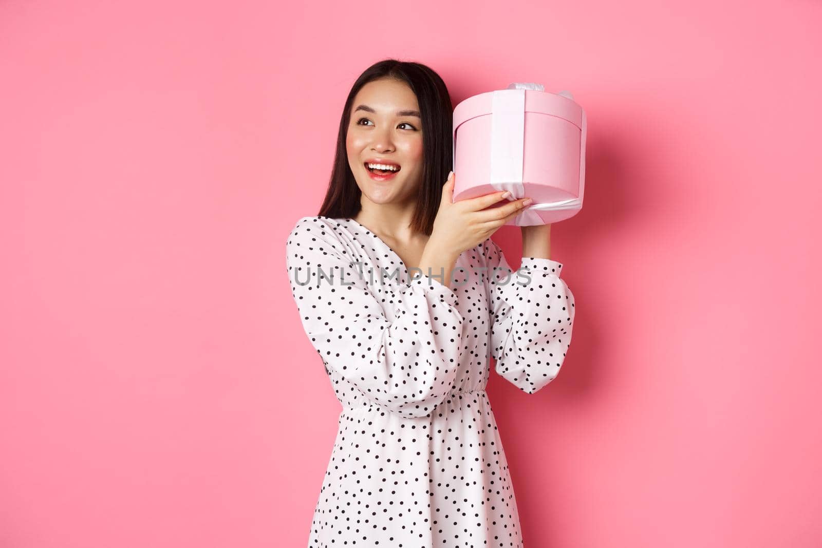 Cute asian woman shaking box with gift, smiling and looking intrigued, guess what inside present, standing over pink background.