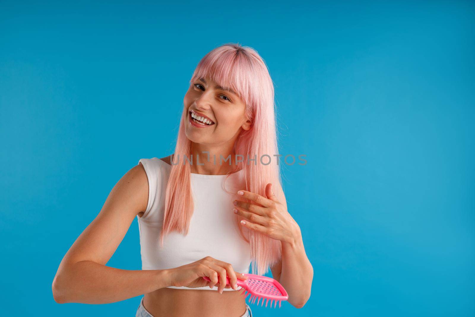 Cheerful woman brushing her smooth natural long pink hair with hair comb while posing isolated over blue studio background. Beauty, hair care concept