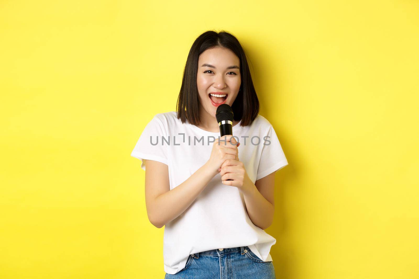 Beautiful asian woman singing karaoke, perform with microphone, smiling happy and looking at camera, standing over yellow background.