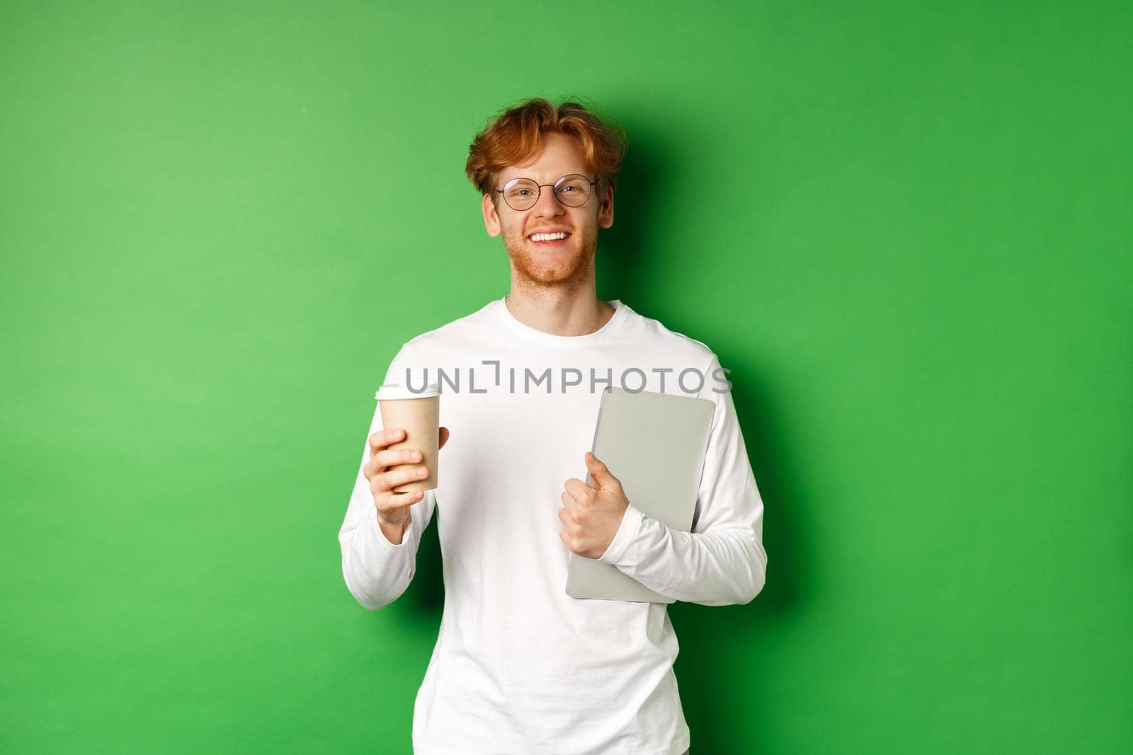 Handsome young man, employee with laptop drinking coffee on break, smiling satisfied, standing over green background.