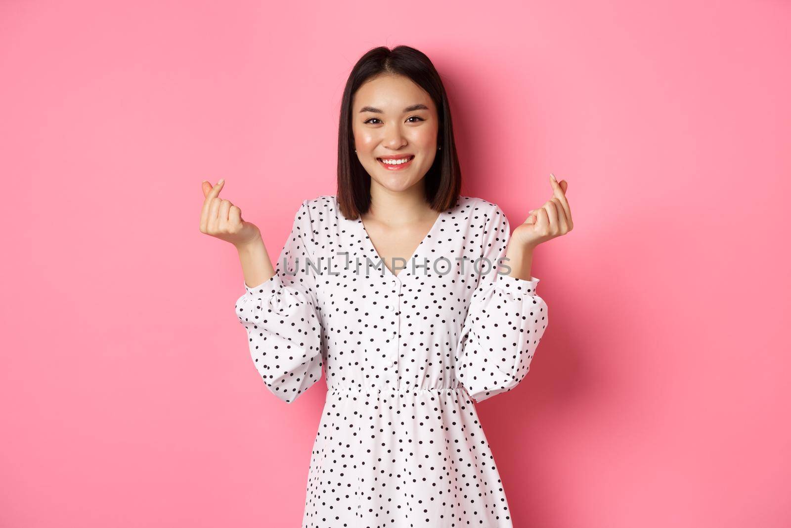 Lovely asian woman in dress showing korean heart signs and smiling, standing on romantic pink background.