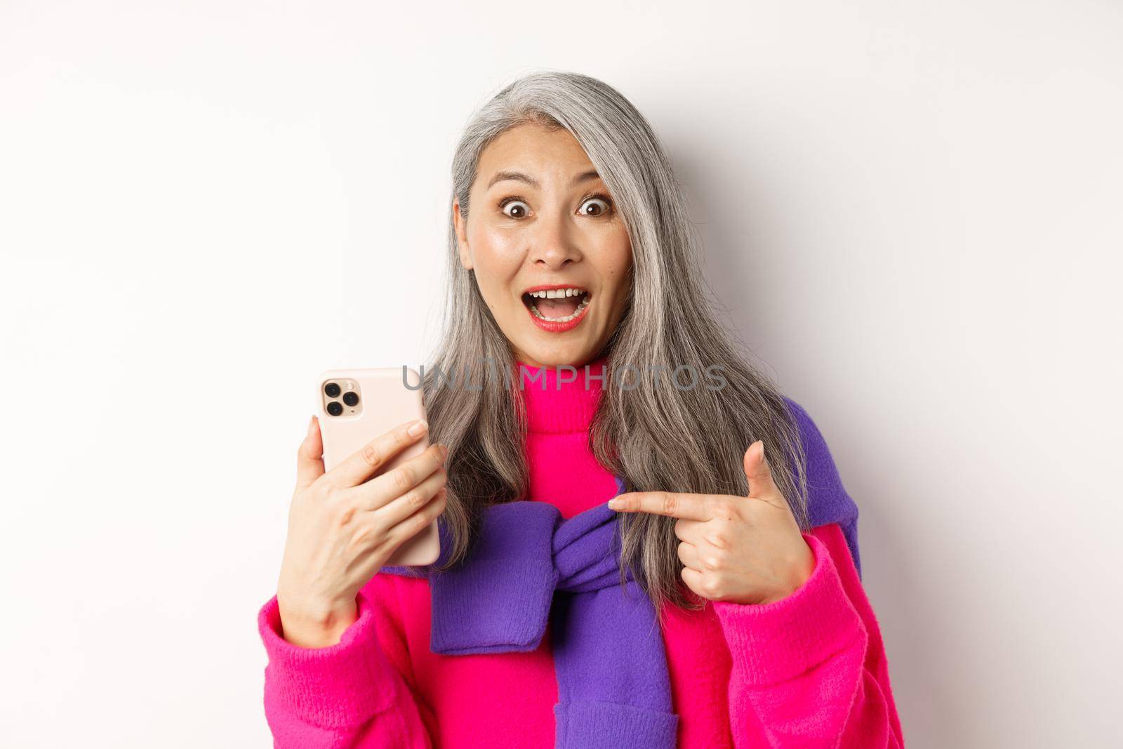 Online shopping. Close up of excited asian senior woman pointing at smartphone and smiling amazed, standing over white background by Benzoix