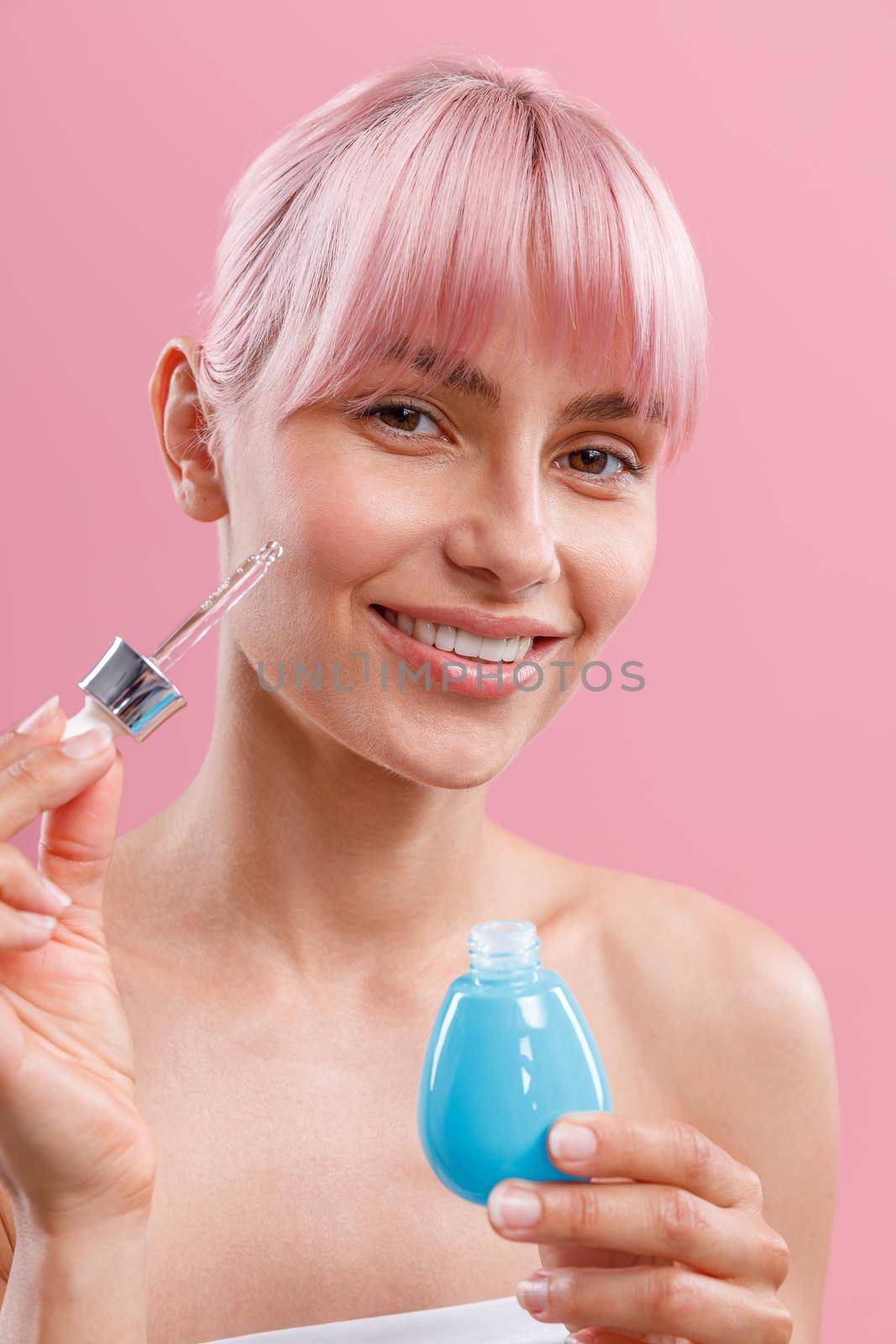 Portrait of happy young woman with pink hair and perfect skin holding dropper and bottle of serum or hyaluronic acid, posing isolated over pink background. Beauty, cosmetics and skincare concept