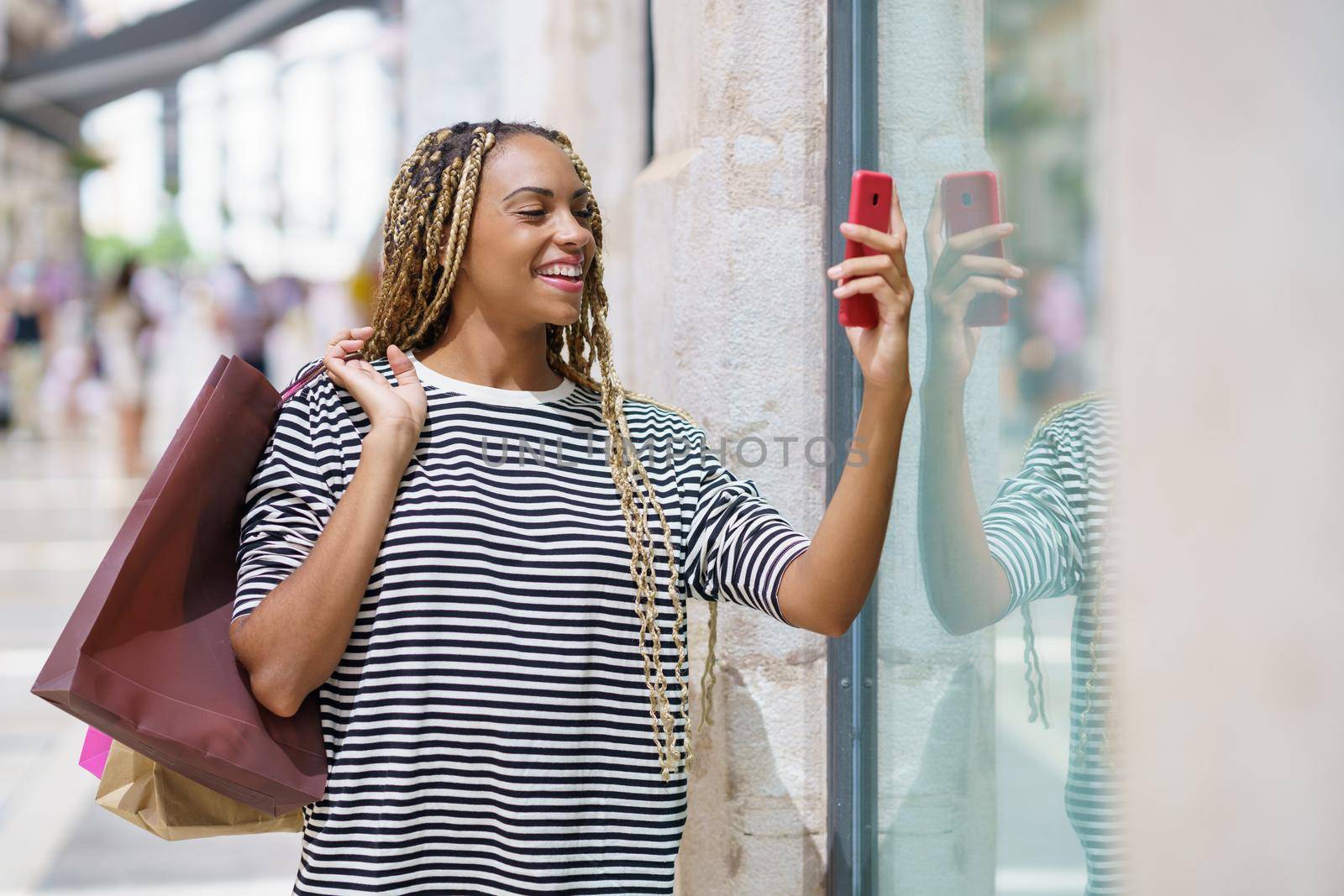 Young black woman photographing a store window in a shopping street with her smartphone. by javiindy