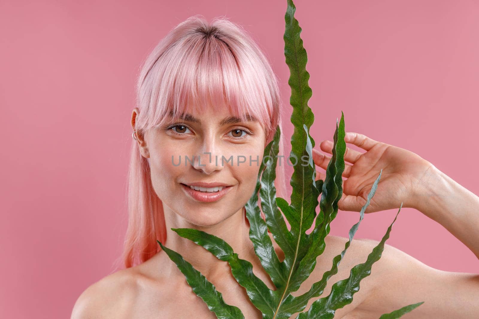 Portrait of beautiful naked woman with pink hair smiling at camera, posing with plant leaf isolated over pink studio background. Beauty, skin care, natural cosmetics concept