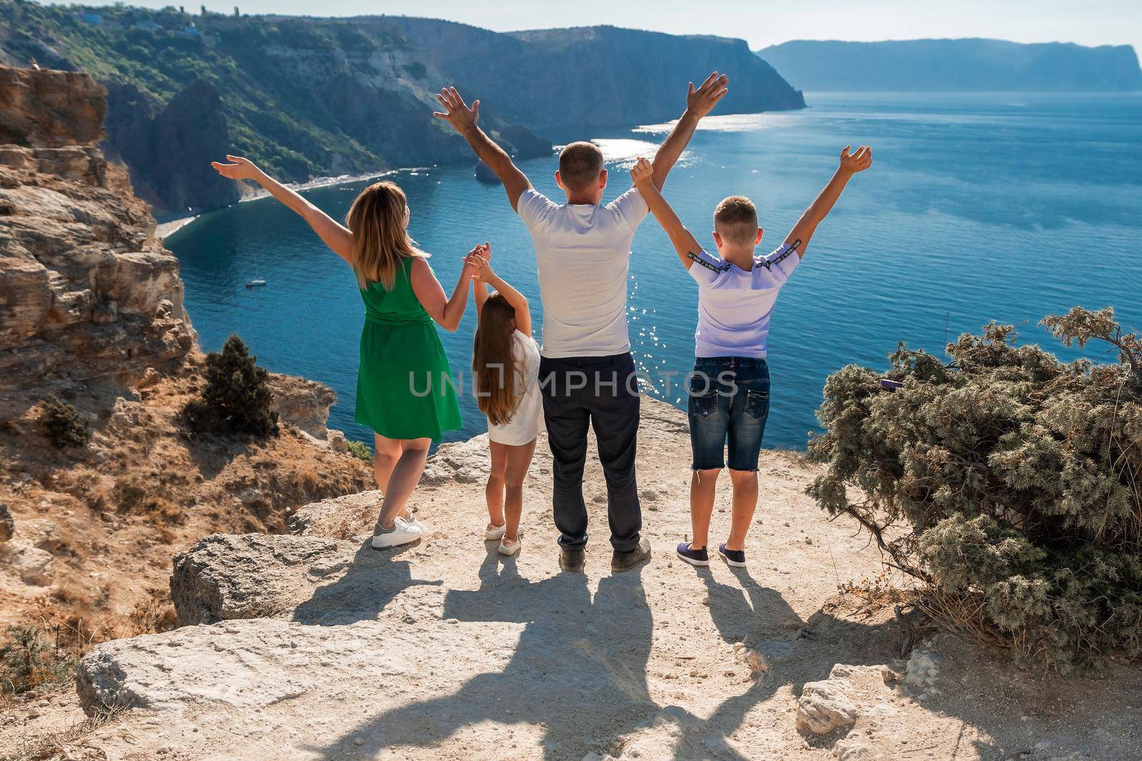 Family mom dad and two children, a boy and a girl stand with their backs to the audience and look out over the sea. They raised their hands. Against the backdrop of a beautiful seashore with rocks