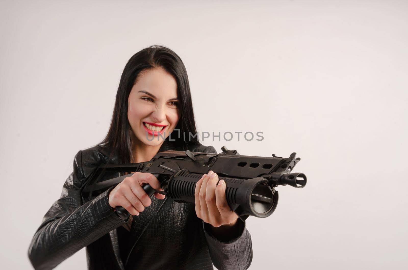 the brunette woman poses in a leather jacket with her back against a white background with a gun