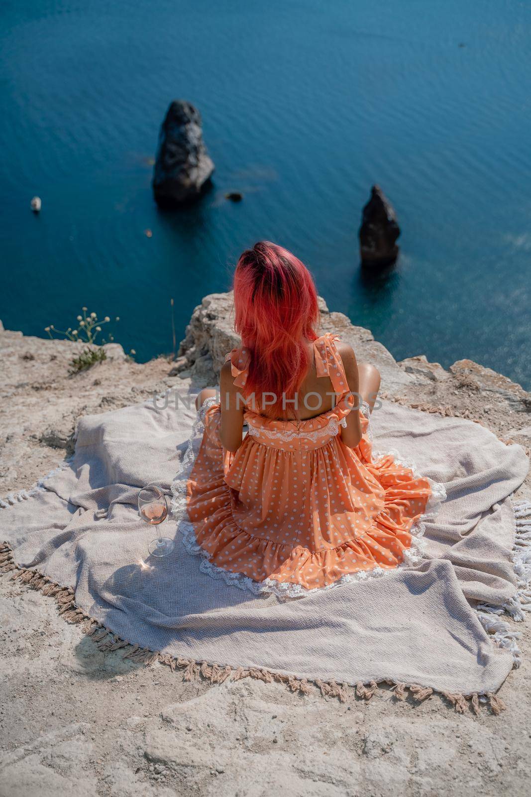 A girl with red hair sits with her back to the viewer on a picnic blanket in an orange dress. Summit on the mountain against the background of the sea and rocks in the sea