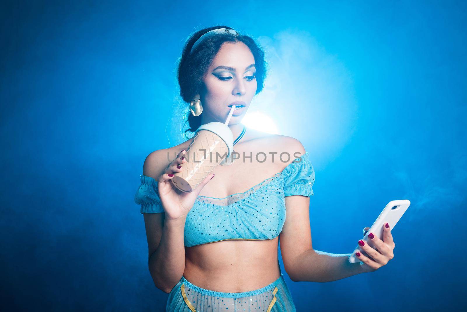 Magic, cosplay and fairy tale concept - Portrait of a young woman in the image of an Eastern fairy Princess holds smartphone and shaker on blue background. by Satura86