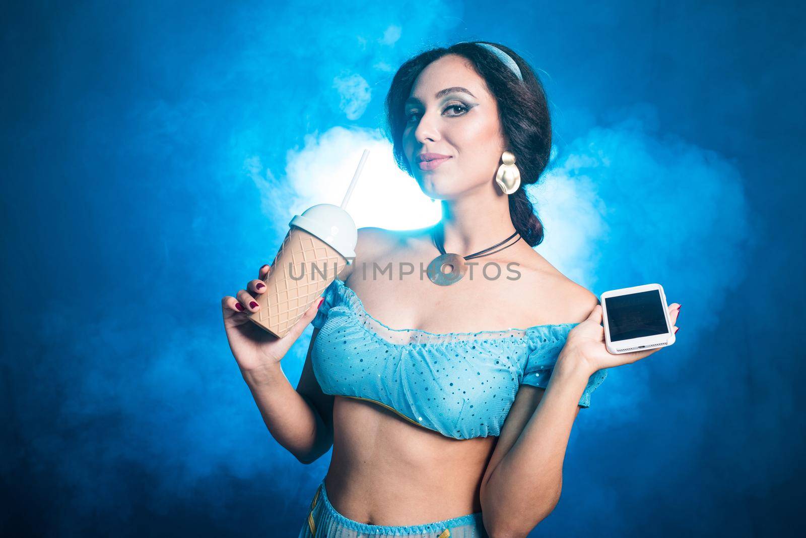 Magic, cosplay, carnival and fairy tale concept - Portrait of a young woman in the image of an Eastern fairy Princess holds smartphone and shaker on blue background. by Satura86