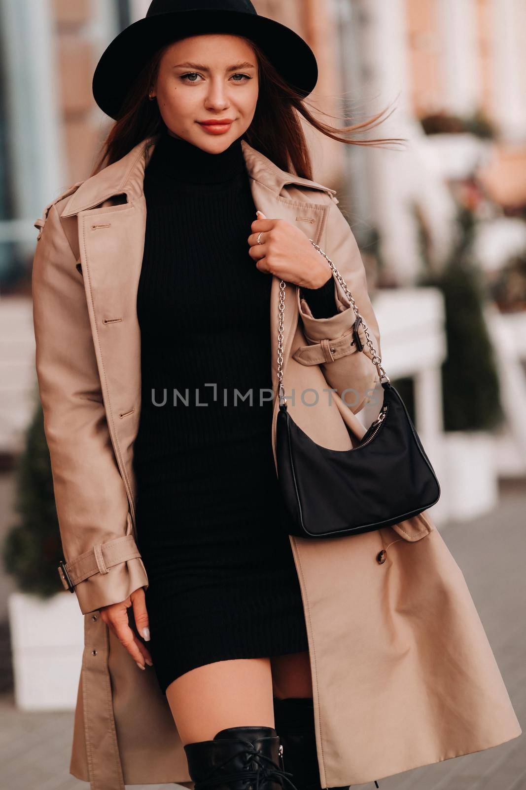 Stylish young woman in a beige coat and black hat and with a black purse on a city street. Women's street fashion. Autumn clothing.Urban style by Lobachad