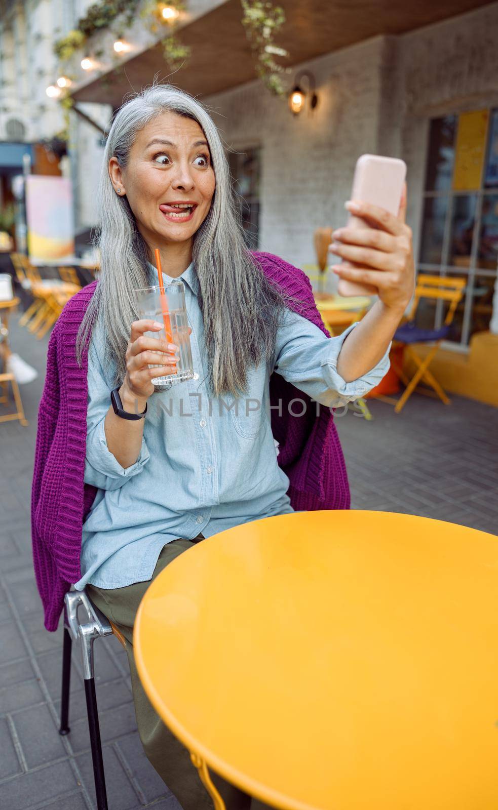 Funny senior Asian woman with glass of water takes selfie with cellphone grimacing at small table on outdoors cafe terrace