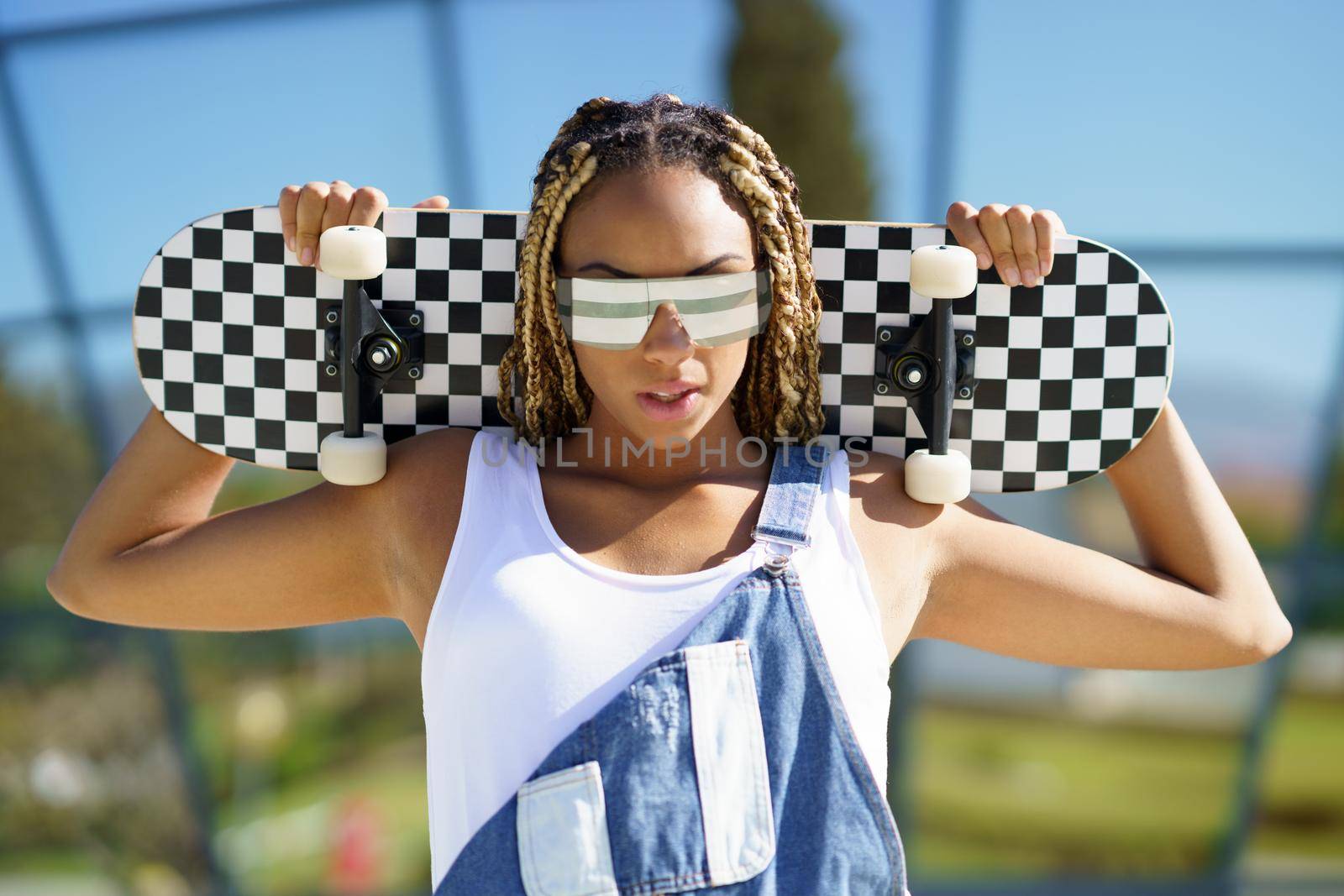 Black young female dressed casual, looking at camera with modern sunglasses and a skateboard.