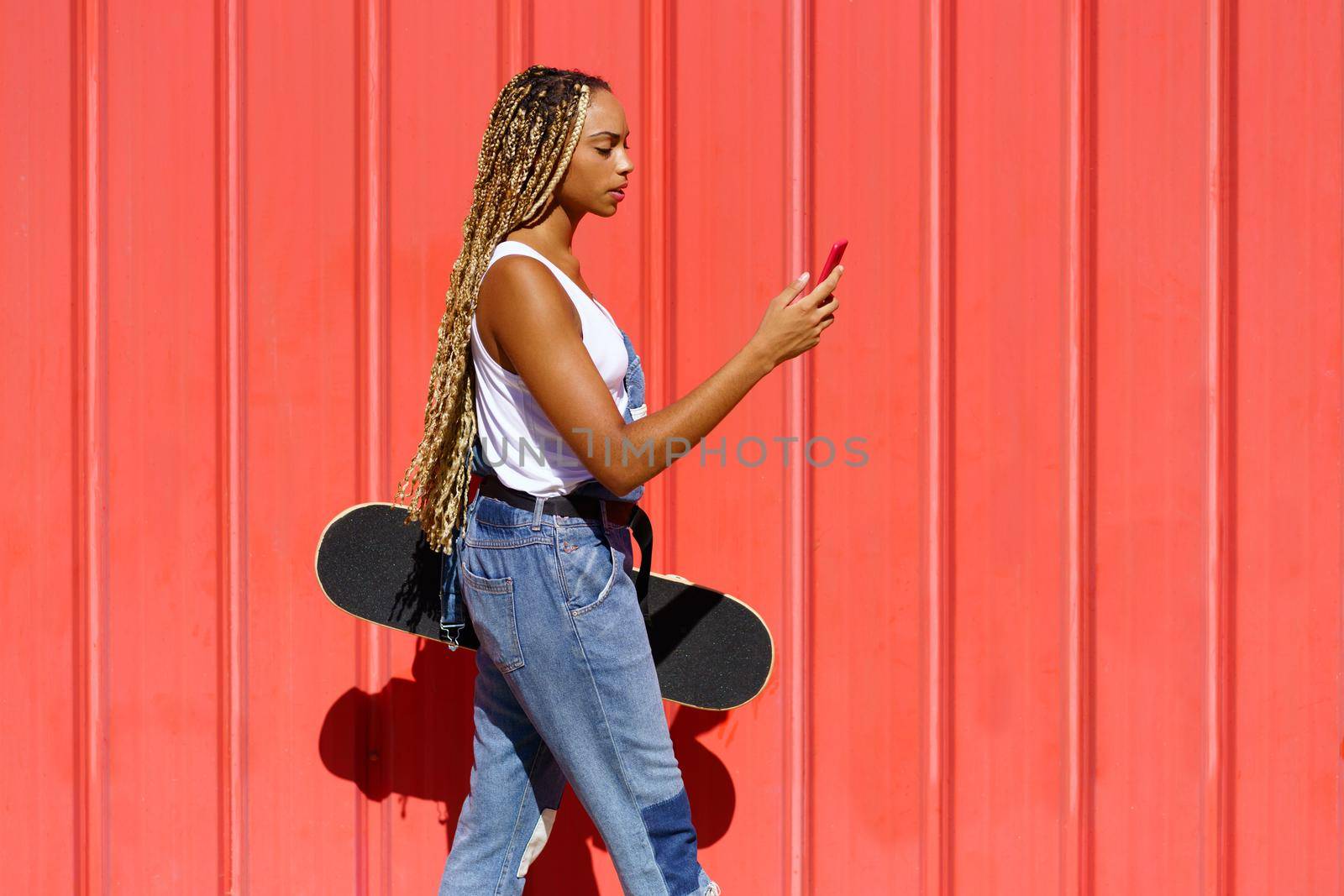 Black woman with coloured braids, consulting her smartphone with her feet resting on a skateboard. by javiindy