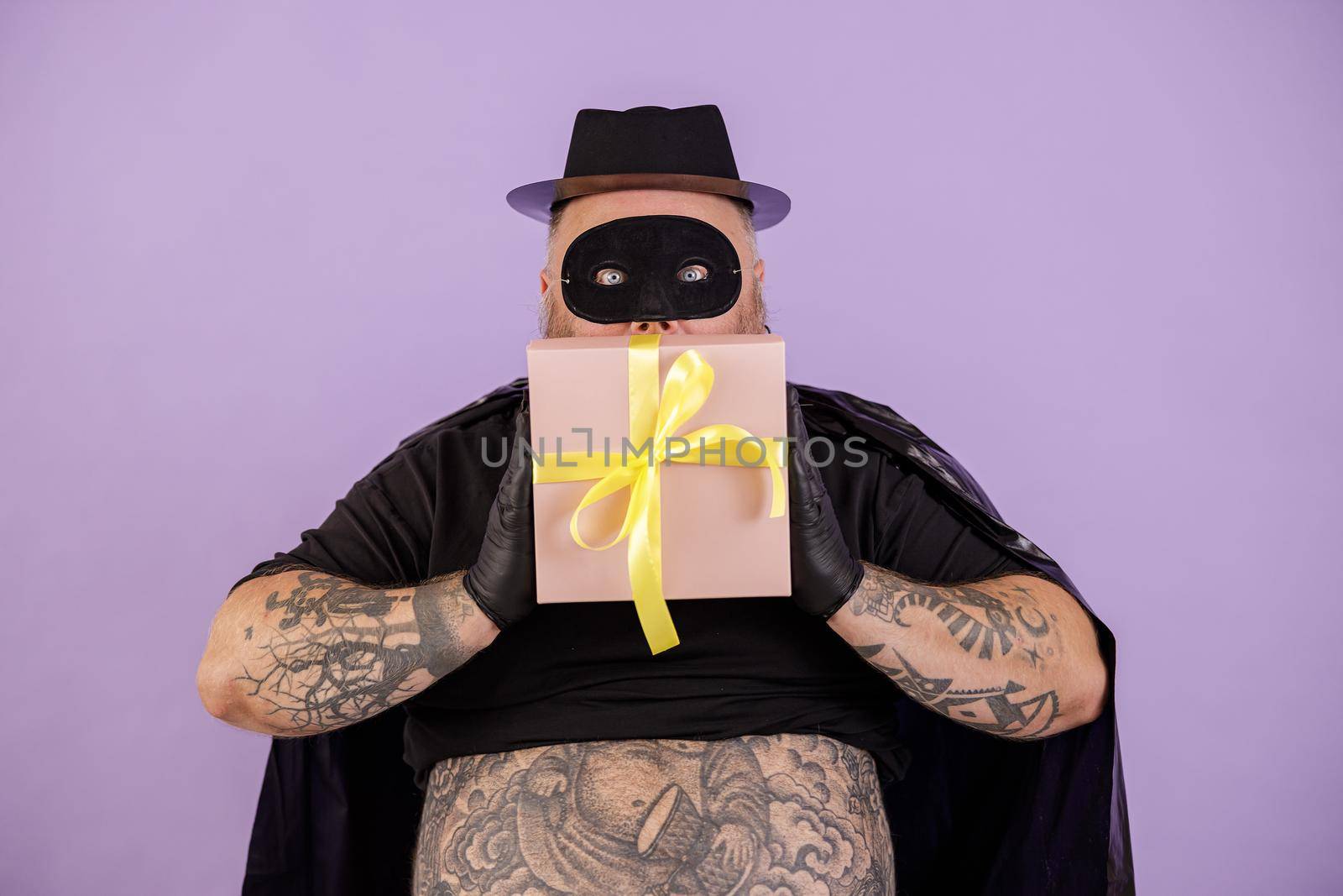 Emotional mature gentleman with overweight and tattoos wearing black hero costume holds present on purple background in studio
