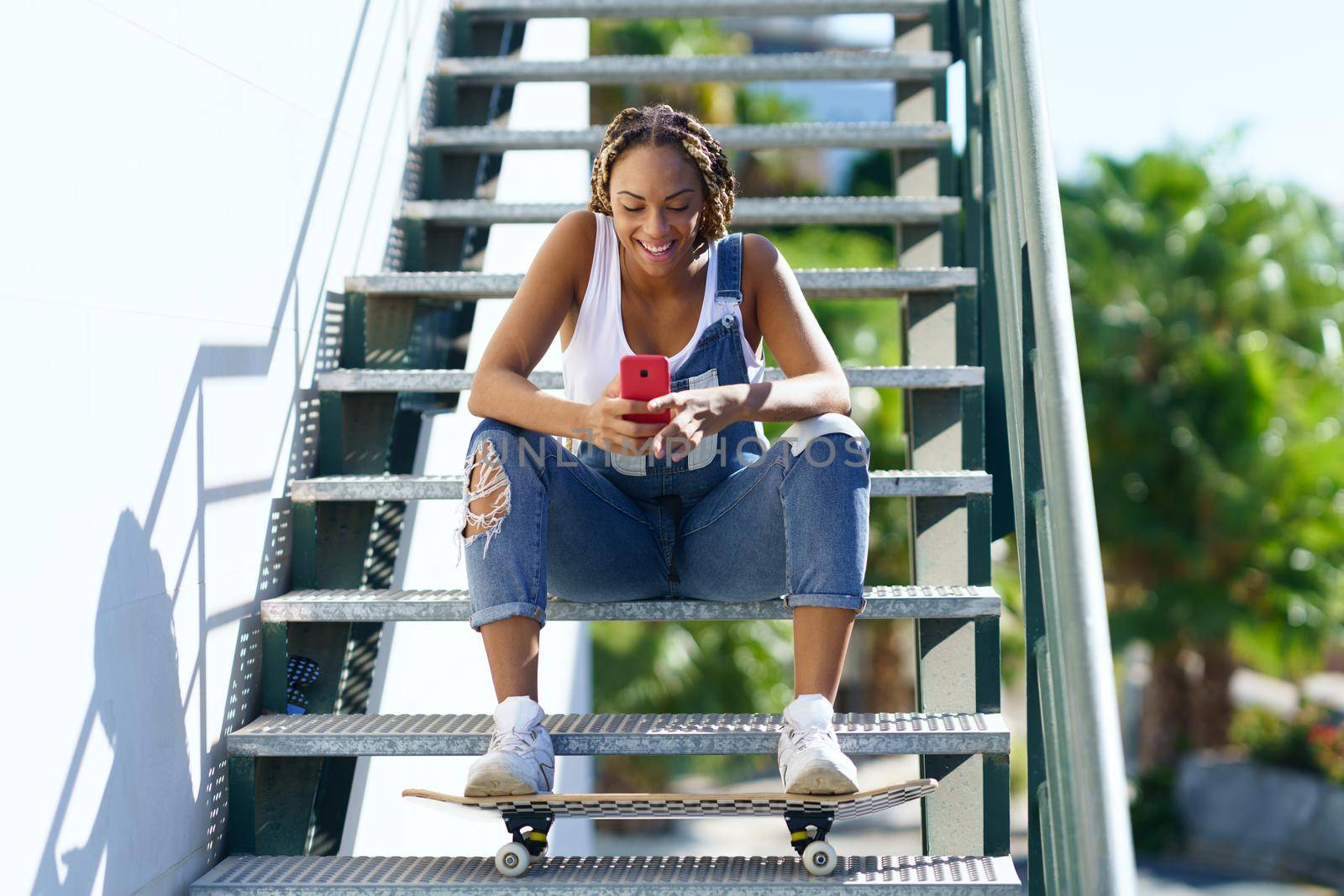 Young black woman with coloured braids, sitting on some steps while consulting her smartphone with her feet resting on a skateboard.