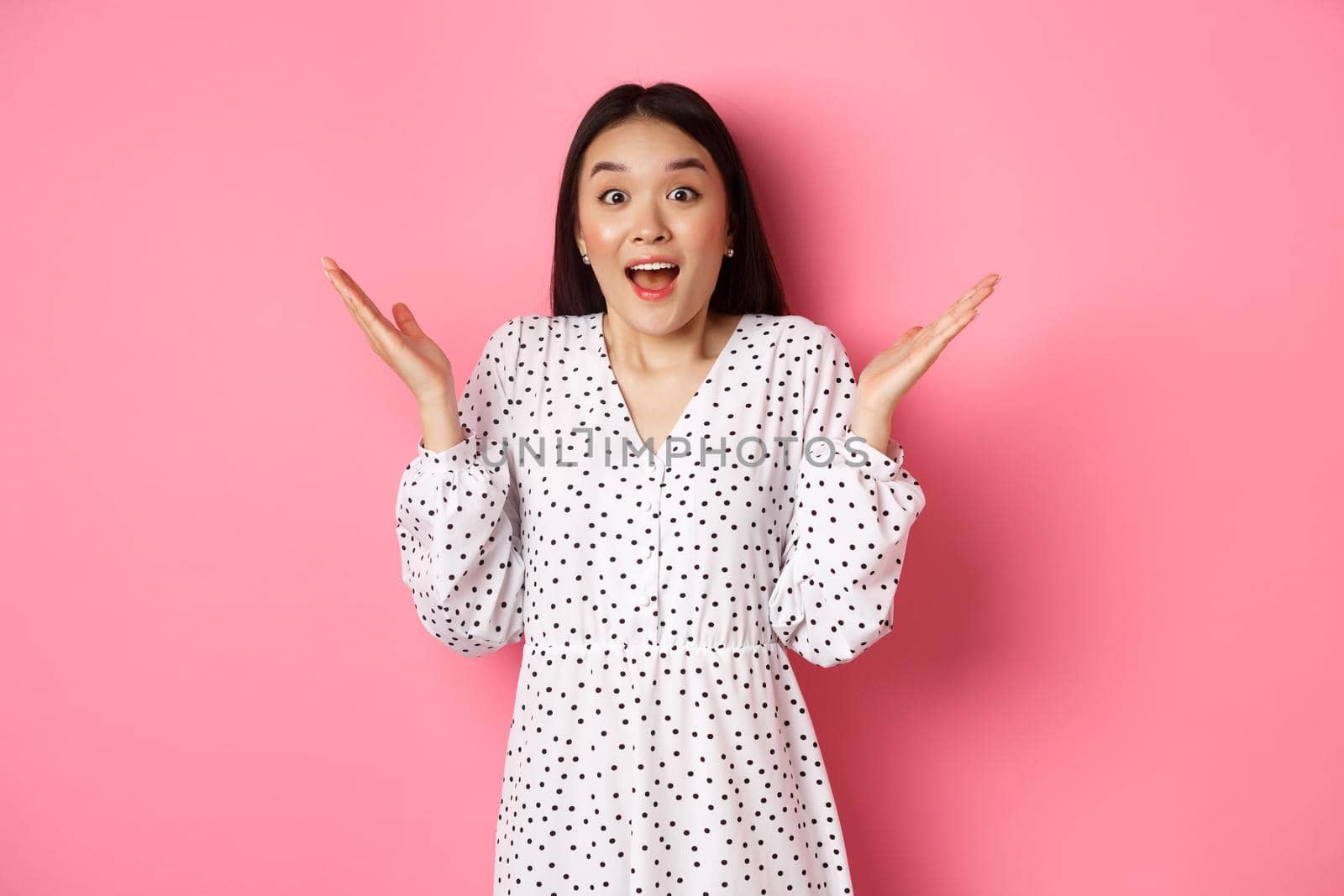 Happy and surprised asian woman rejoicing, spread hands and gasping amazed, looking with excitement and disbelief, standing over pink background by Benzoix