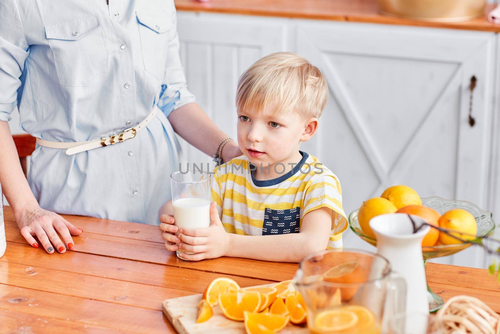 The boy drinks milk from a glass. Mother and son are smiling while having a breakfast in kitchen. Mom is pouring milk into glass by Malkovkosta