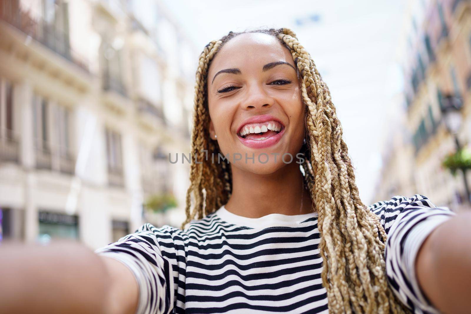 Black woman with afro braids taking a smiling selfie in an urban street with a smartphone. by javiindy