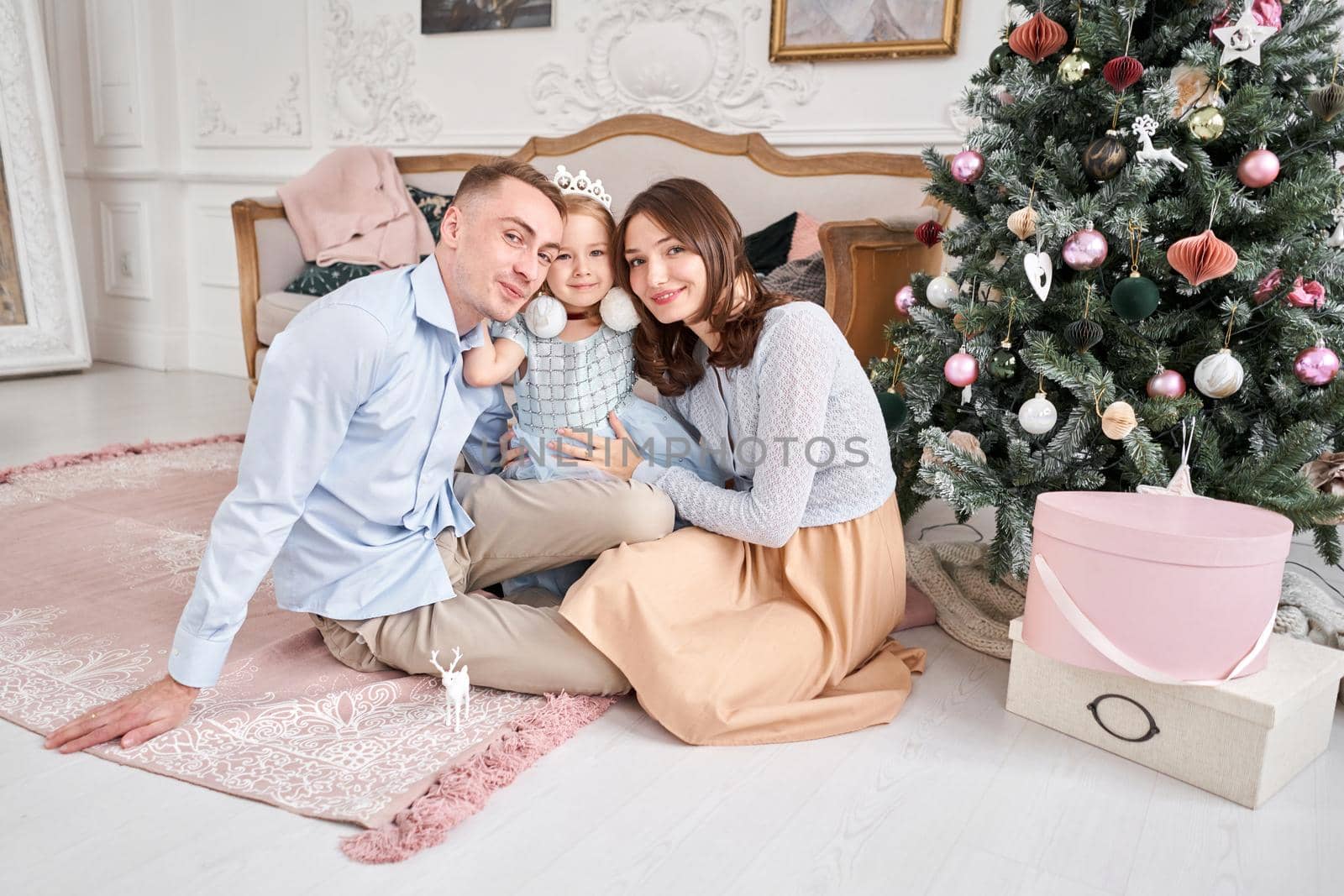 Loving family. Mom and dad hugging little daughter . Parents and baby child having fun near Christmas tree and white fireplace indoors. Merry Christmas and Happy New Year. Cheerful pretty people