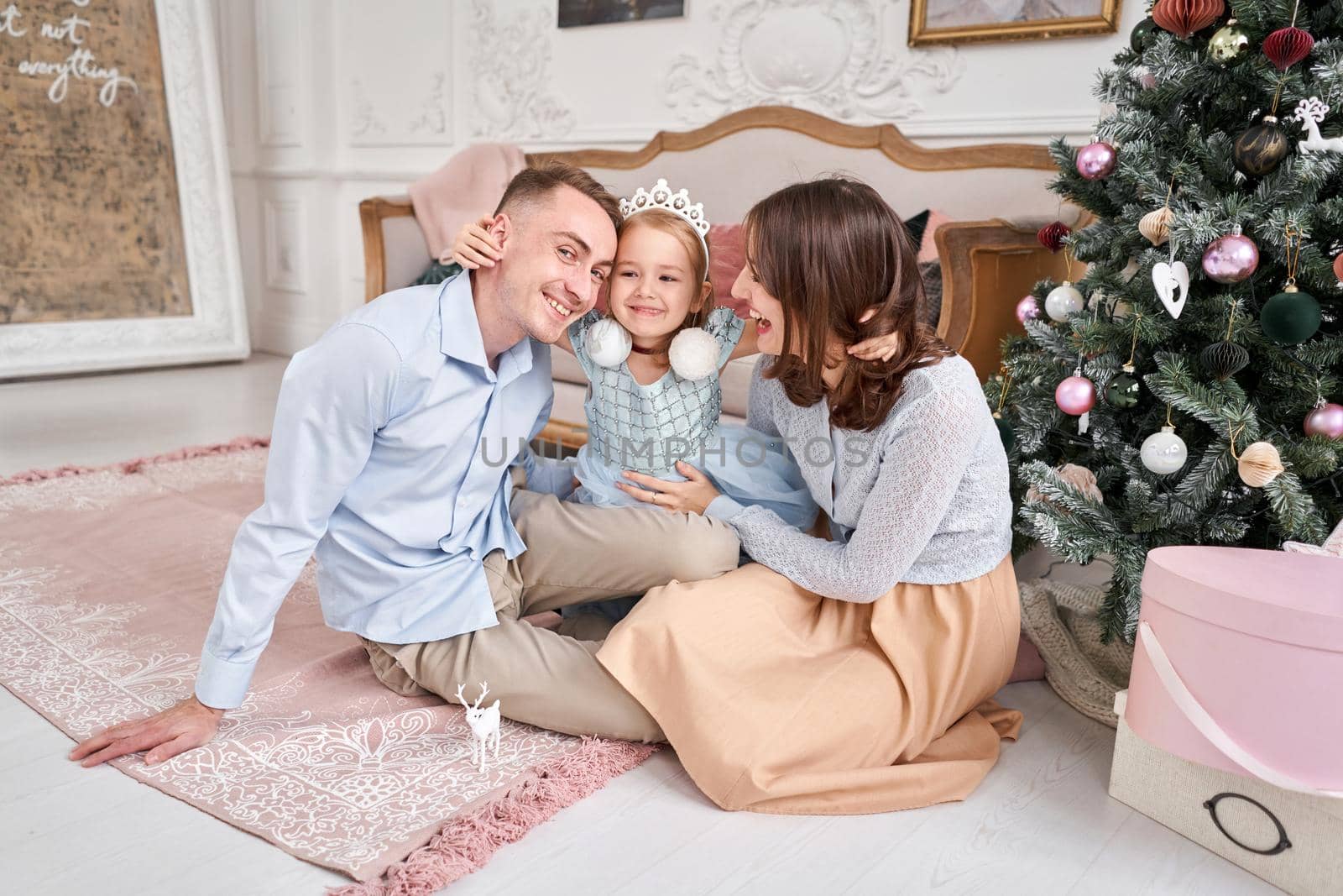 Loving family. Mom and dad hugging little daughter . Parents and baby child having fun near Christmas tree and white fireplace indoors. Merry Christmas and Happy New Year. Cheerful pretty people