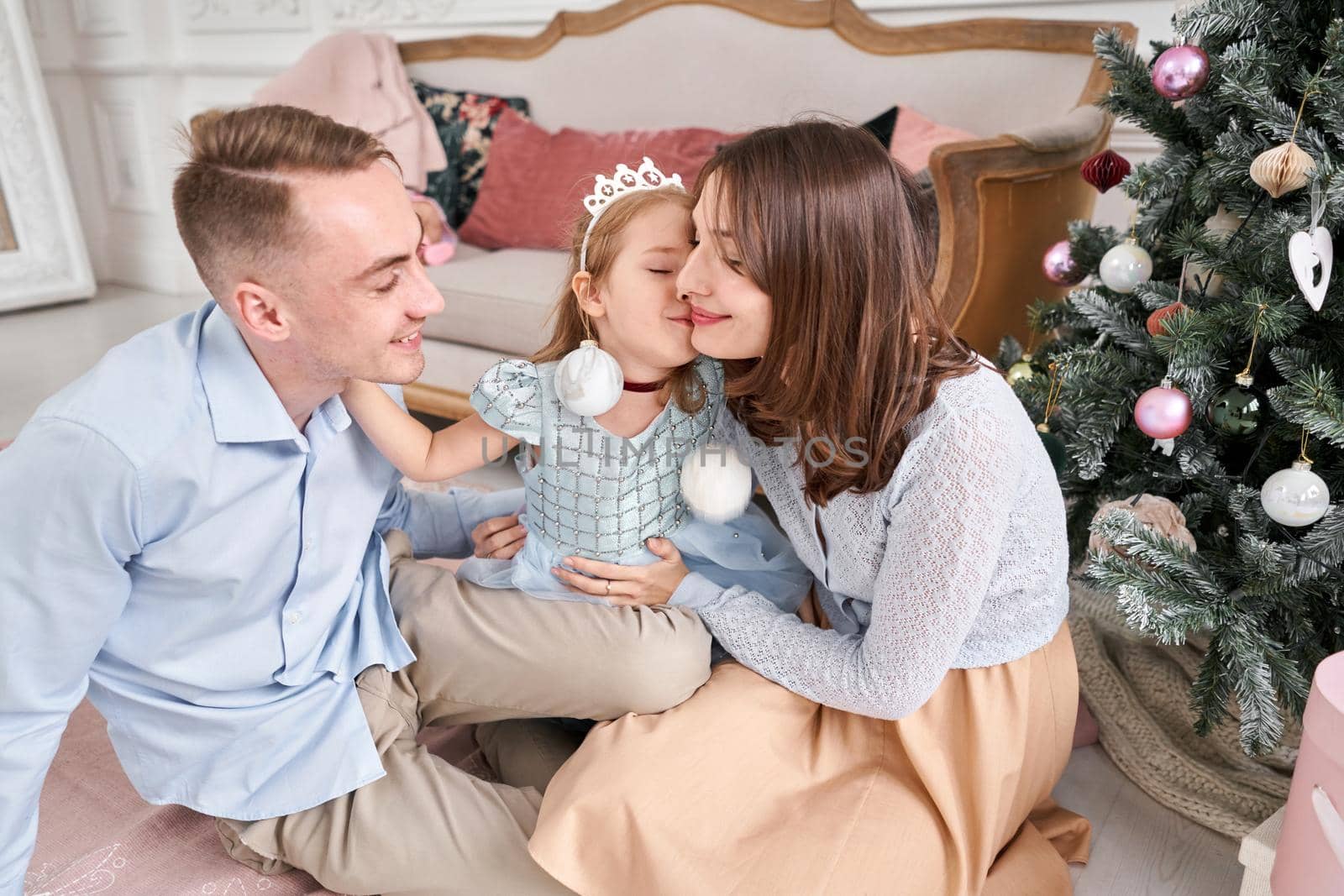 Loving family. Mom and dad hugging little daughter . Parents and baby child having fun near Christmas tree and white fireplace indoors. Merry Christmas and Happy New Year. Cheerful pretty people