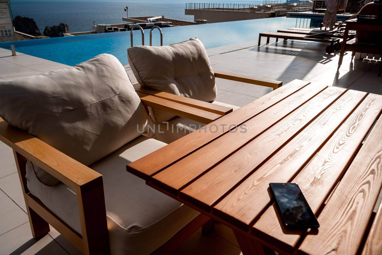 Restaurant with sea view. You can see rocks in the sea. Wooden tables and chairs with white cushions. Overcast, cloudy weather. by Matiunina