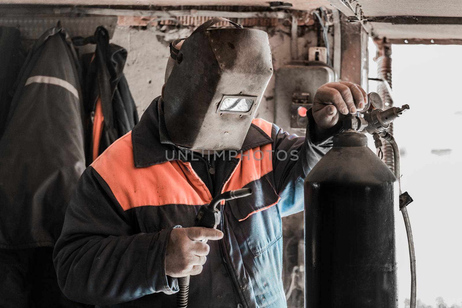 A man welder worker stands in front of a welding machine and a carbon dioxide cylinder, with a gearbox in the shop of an industrial plant.