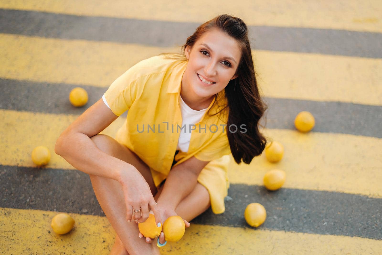 a girl with lemons in a yellow shirt, shorts and black shoes sits on a yellow pedestrian crossing in the city. The lemon mood by Lobachad