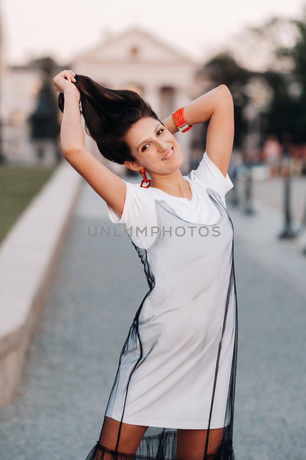 A girl with long hair with red earrings in white clothes walks around the city.The model holds her hair in her hands and smiles.