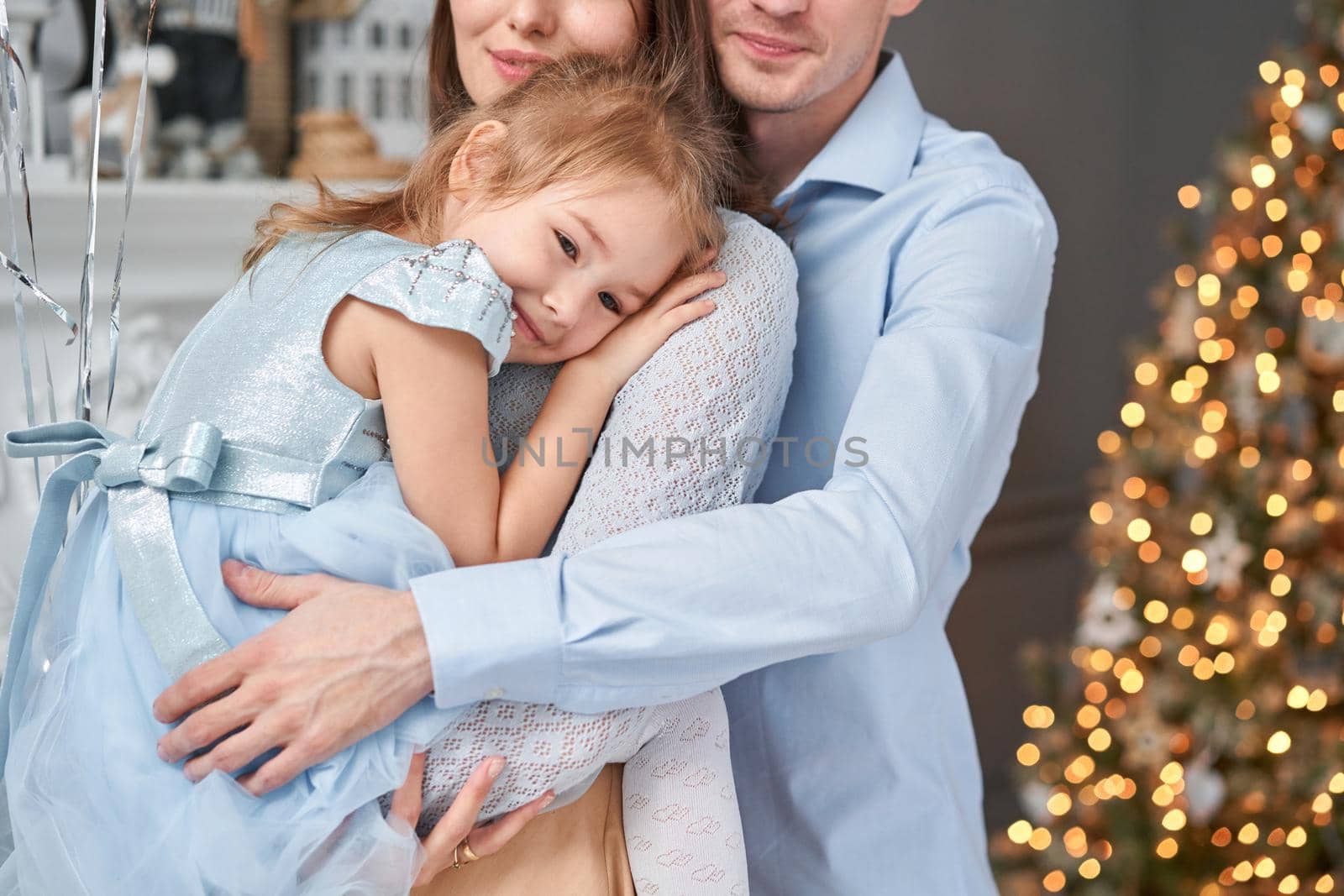 Loving family. Mom and dad hugging little daughter . Parents and baby child having fun near Christmas tree and white fireplace indoors. Merry Christmas and Happy New Year. Cheerful pretty people