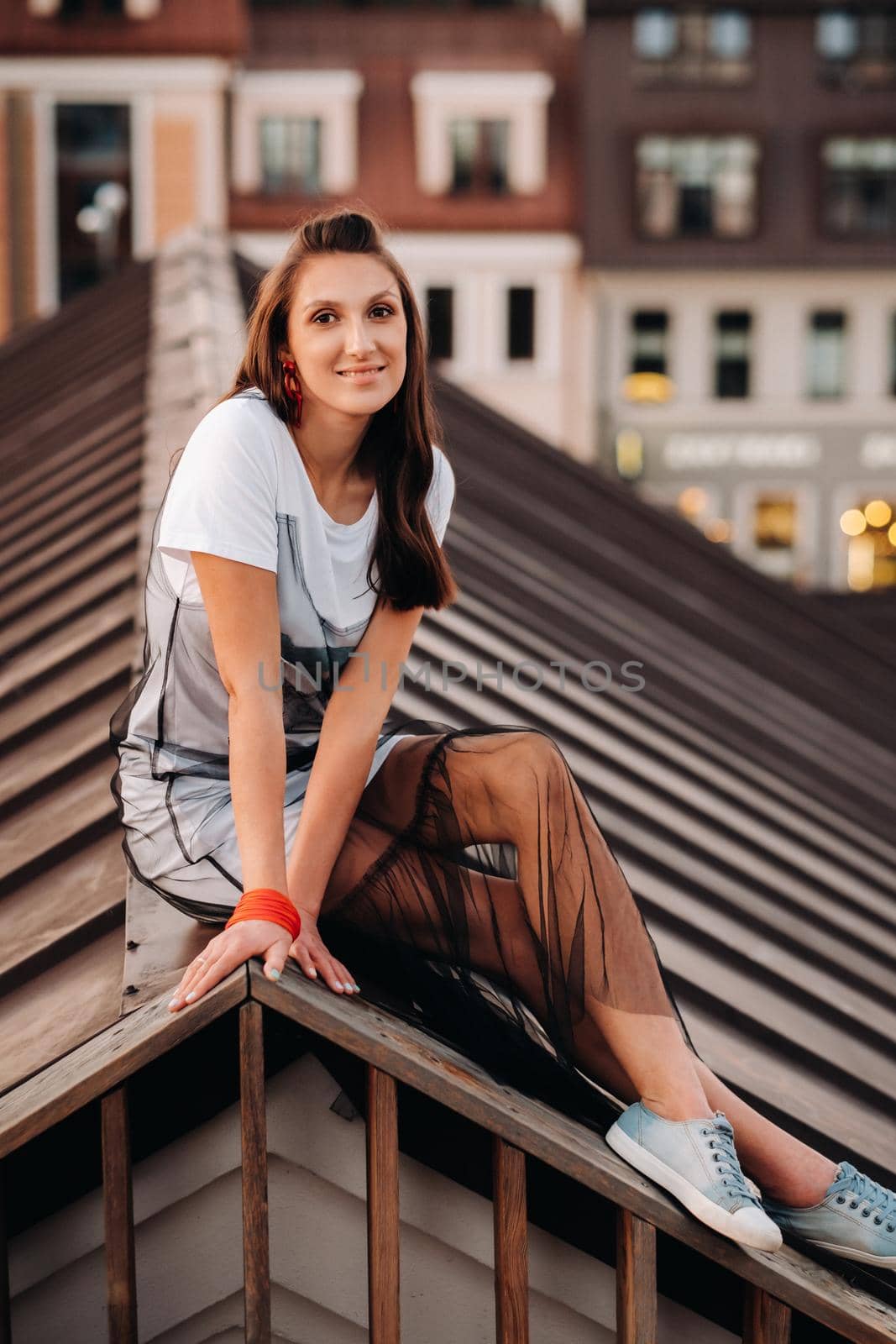 A girl sits on the roof of a house in the city in the evening. Portrait of a model in a dress and sneakers. by Lobachad