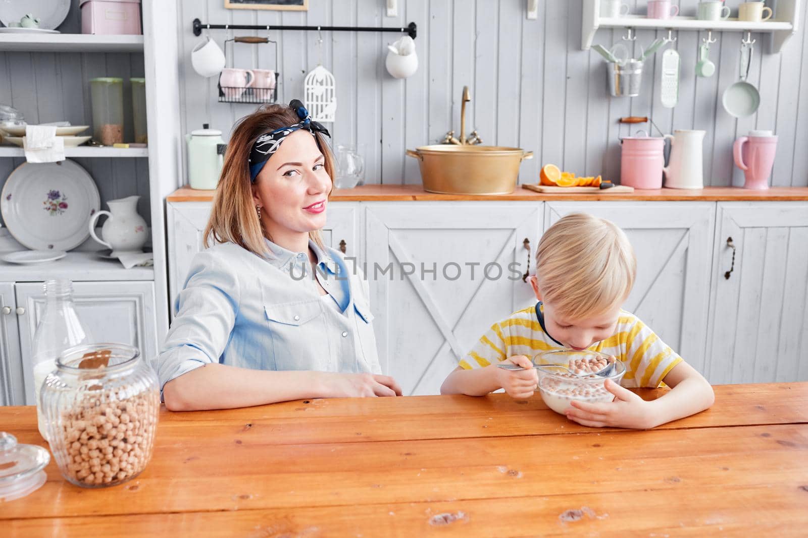 The boy eats cereal with milk. Mother and son are smiling while having a breakfast in kitchen. Bright morning in the kitchen. Healthy Breakfast cereals and fresh fruit. by Malkovkosta