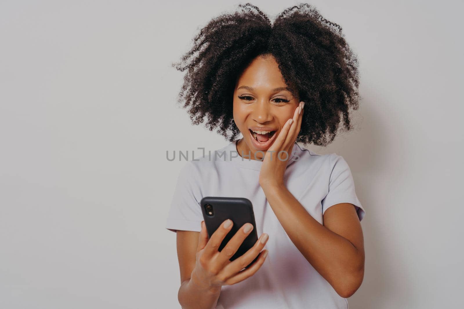 Excited mixed race ethnicity millennial girl isolated on white studio background feeling delightfully winning on cellphone app, overjoyed happy african young woman gets great news on smartphone