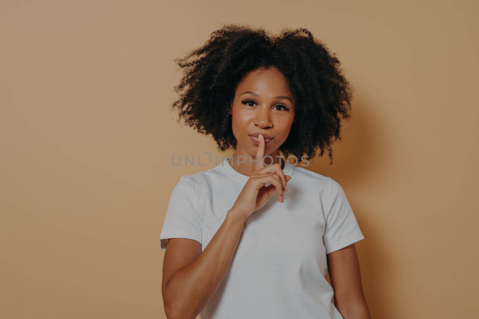 Close up portrait of afro american attractive mysterious wavy-haired woman in white tshirt showing shh sign with finger, isolated on pastel yellow color background with copy space for advertisement