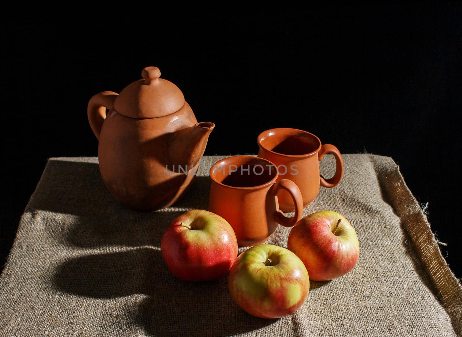 still life with a clay teapot, two cups and three red apples. indoor closeup on black background