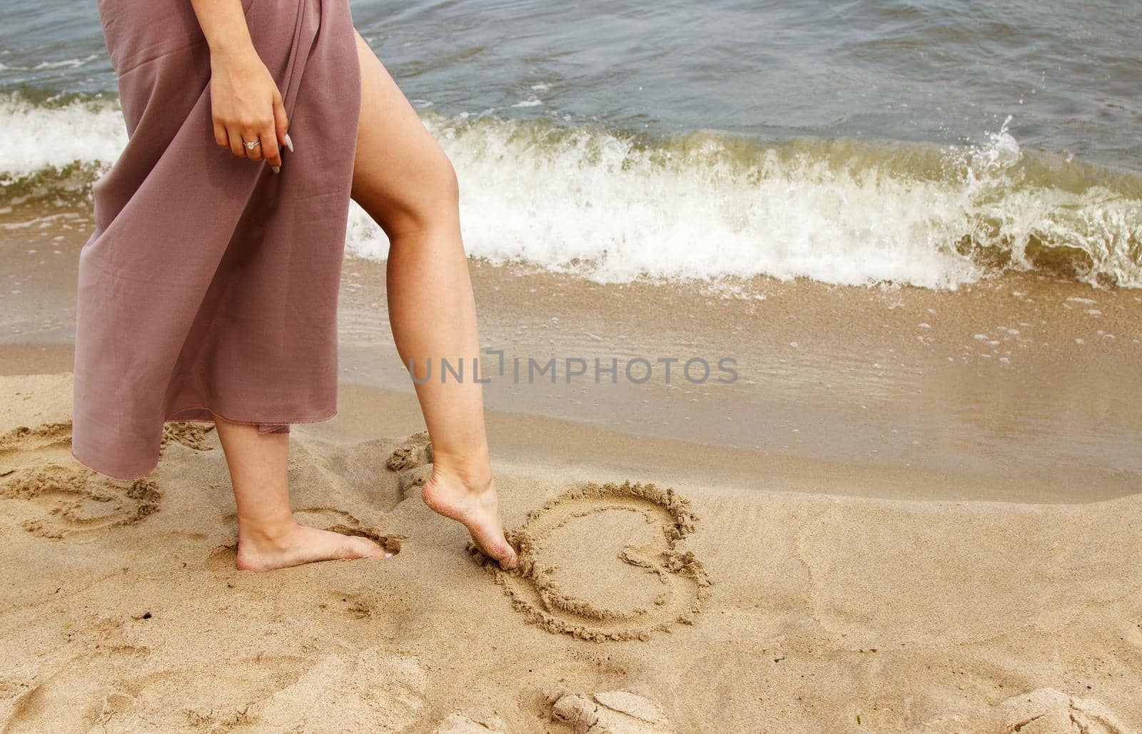 woman in beige dress draws with her foot heart on the sand by sea on summer day. outdoor closeup