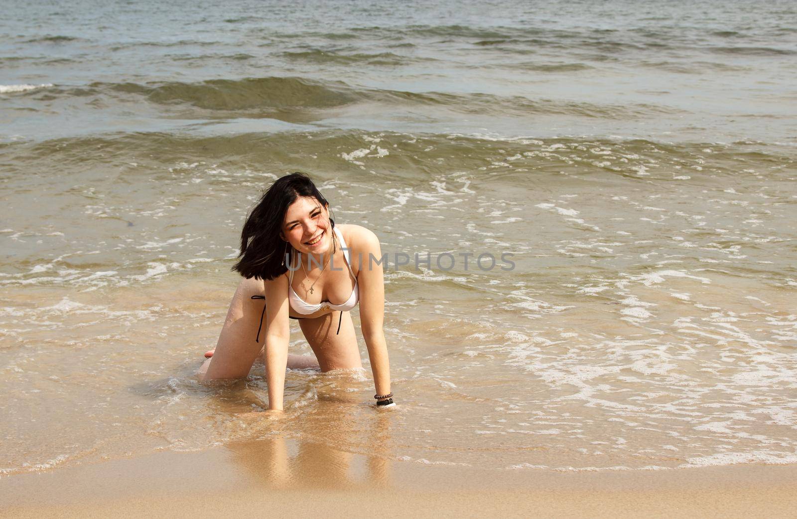 young brunette woman in bikini posing kneeling on the sand by the sea on sunny summer day