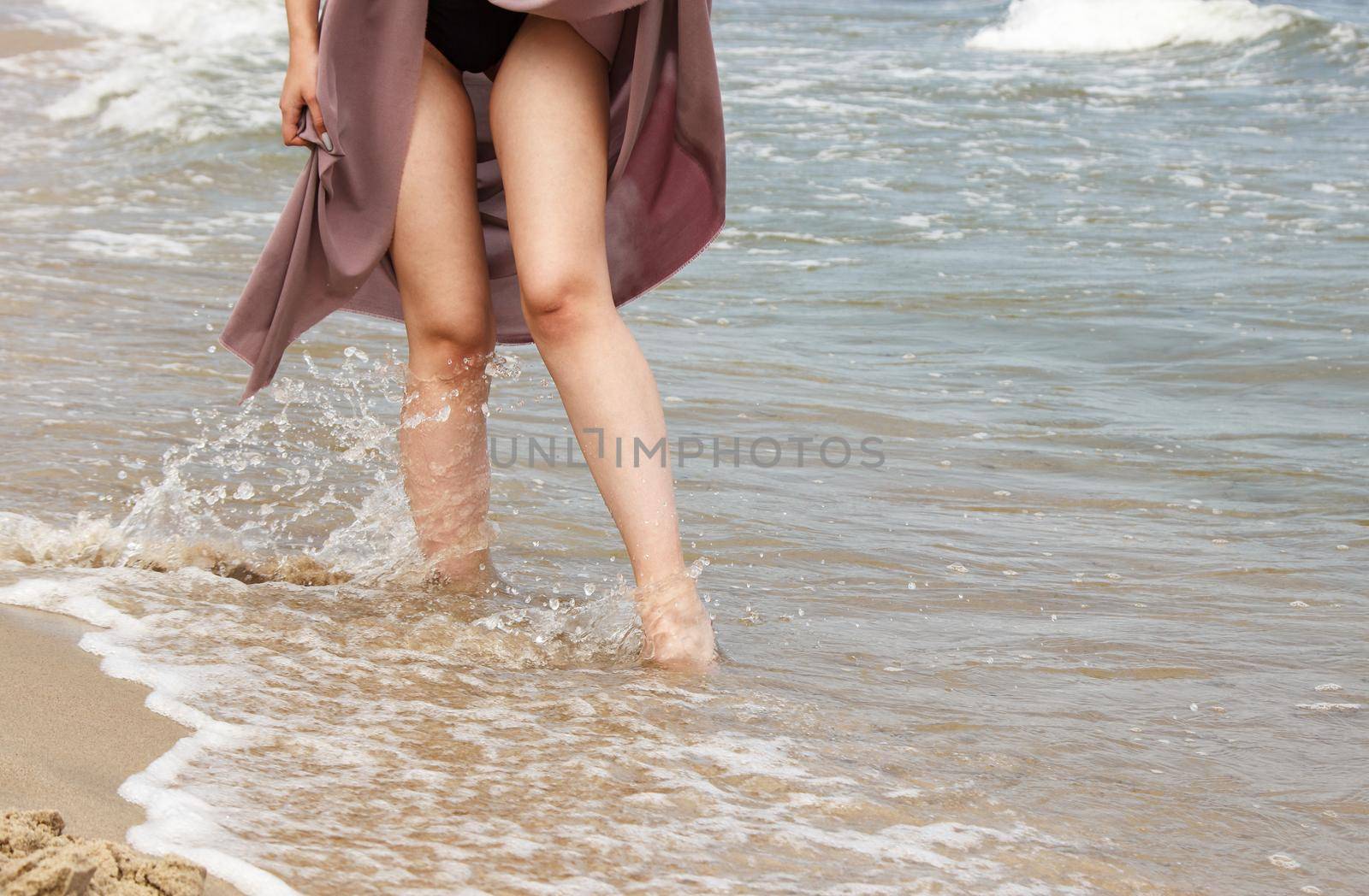 legs of a young woman in black bikini and beige dress standing on sand by sea by raddnatt