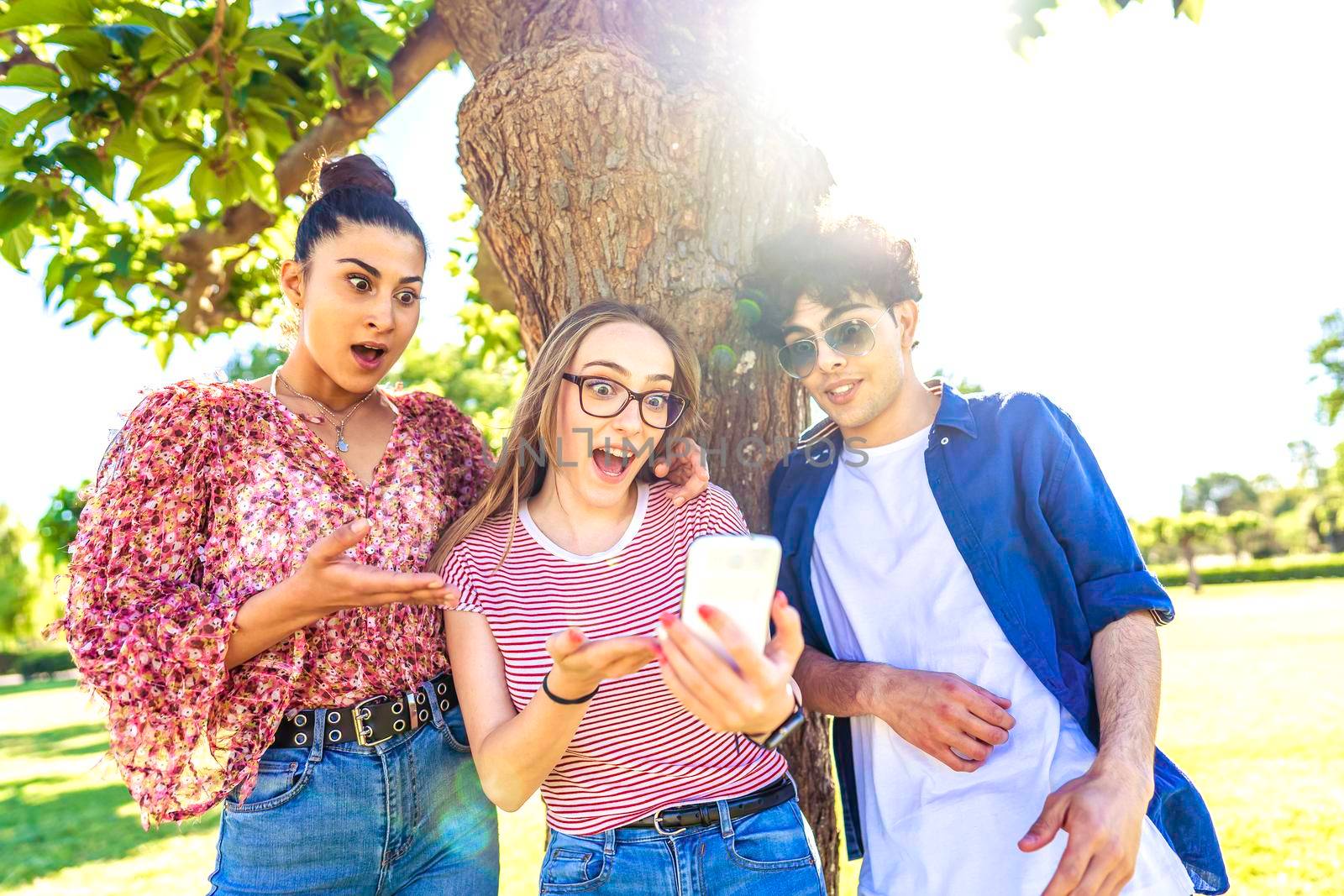 Three diverse students making surprised faces spreading mouth and eyes pointing and looking smartphone spending time in nature of city park. power of new wifi technology addicting all ages people by robbyfontanesi